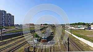 An electric locomotive stands on graveled and electrified railroad tracks. Behind a concrete fence are residential houses and a ci