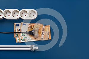 Electric plug on 50 euro banknotes, power strip and compact fluorescent light bulb over blue background. High energy costs concept