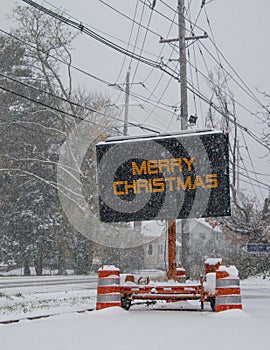 Electric road traffic mobile sign by the side of a snow covered road with snow falling that says, Merry Christmas.