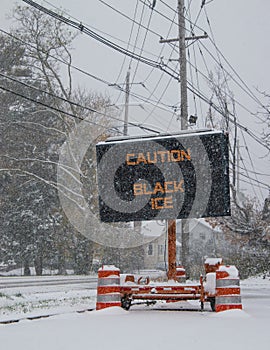 Electric road traffic mobile sign by the side of a snow covered road with snow falling warning of black ice on road.