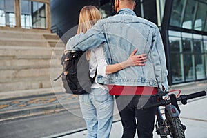 With electric schooter. Young stylish man with woman in casual clothes outdoors near business building together. Conception of