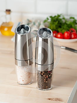 Electric spice mills made of metal. On a light wooden background. In the background is a plate with vegetables and olive oil