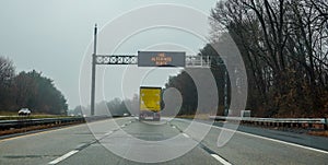 Electric warning sign over yellow box truck on highway advising drivers to use an alternate route on an overcast day.