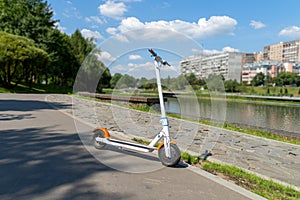 An electric white scooter stands on the bandwagon on the street. City park along the promenade. Sunny summer day.