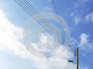 Electric wooden pole with lamp and electricity cables against blue sky and white clouds.