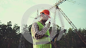 Electrical equipment worker near high voltage tower using walkie talkie and clipboard. Industrial engineer and power