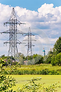 Electrical supports of a high-voltage line. Green trees and high-voltage power towers against the background of an old Church