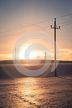 Electrical tower on a snowy field. Sun goes down, orange colors, evening scenery