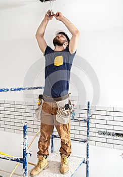 Electrician Builder at work, installation of lamps at height. Professional in overalls with an electrical tool. On the background