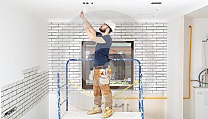 Electrician Builder at work, installation of lamps at height. Professional in overalls with an electrical tool. On the background