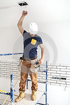 Electrician Builder at work, installation of lamps at height. Professional in overalls with an electrical tool. On the background