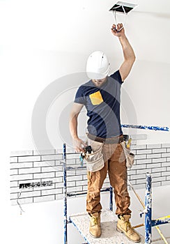 Electrician Builder at work, installation of lamps at height. Professional in overalls with an electrical tool. On the background