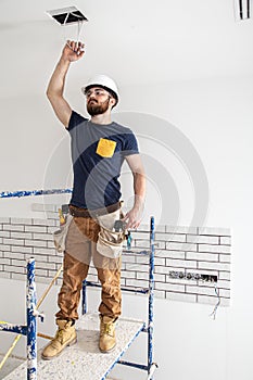 Electrician Builder at work, installation of lamps at height. Professional in overalls with an electrical tool. On the background