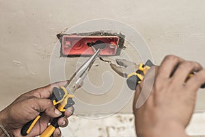 An electrician handling an exposed wire in an outlet with a pair of pliers. Replacing a faulty electrical outlet at a living room