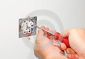 Electrician installing a grey wall-mounted AC power socket with a screwdriver on a white wall, renovating home.