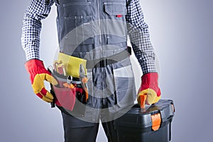 Electrician in jumpsuit with toolbox against gray background.