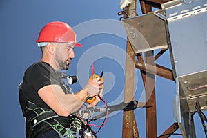 Electrician in red helmet and safety belt with multimeter on the electric pole