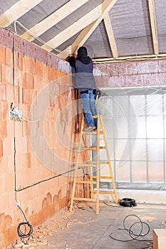 Electrician standing on ladder installing cables in the channel / groove along with electrical boxes in unfinished house.