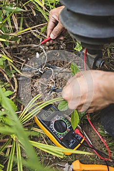 An electrician tests an exposed grounded wire with a digital multimeter with LCD screen. Safety procedure at a home garden