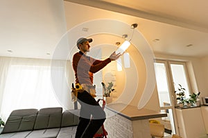 Electrician in uniform repairing ceiling lamp indoors