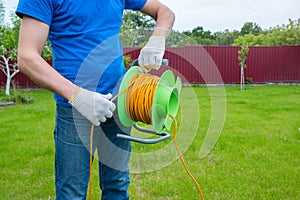 An electrician is unrolling an extension cord on the coil in the house backyard. Cleaning of the territory