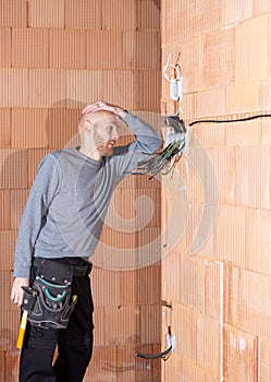 An electrician at work thinking what start to install first in sockets in unfinished house built of clay block bricks.