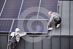 Electricians connecting cables while installing photovoltaic solar panels on roof of house.