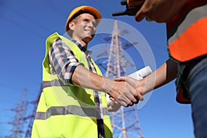 Electricians shaking hands near high voltage tower, closeup