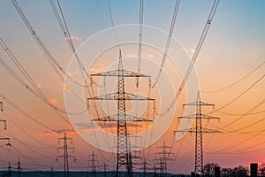 Electricity pylons and electric power transmission lines against vibrant orange sky at sunset. High Voltage towers provide power.