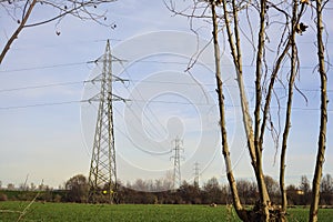 Electricity pylons and over head cables in the middle of a field framed by bare trees in the italian countryside