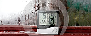 Electronic clock, calendar, thermometer, and hygrometer, against the background of condensation on glass, high humidity. Digital