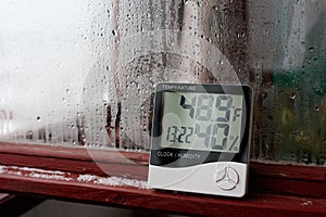 Electronic clock, calendar, thermometer, and hygrometer, against the background of condensation on glass, high humidity. Digital