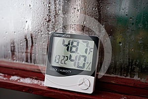 Electronic clock, calendar, thermometer, and hygrometer, against the background of condensation on glass, high humidity. Digital