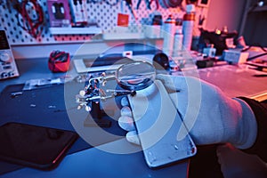 Electronic technician holds a modern smartphone with a broken body, carefully examines the damage using a magnifying