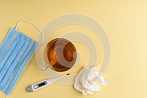 Electronic thermometer, cup of tea, mask and green pills on yellow background.