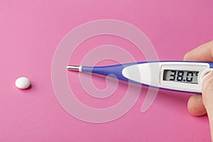 Electronic thermometer and pill on a pink background. High temperature 38 degrees Celsius on display