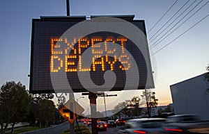 Electronic traffic sign stating Expect Delays with blurred traffic at sunset