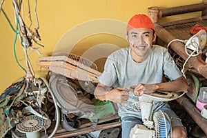 Electronics repairman smiles at the camera while holding the fan cable while sitting around broken items