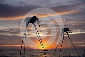 Elefanten auf Stelzen als Touristenattraktion am Sunset Sanato Beach Club bei Sonnenuntergang mit Fischerboot im Hintergrund in