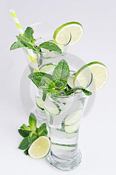 Elegance glasses with cold natural cocktail with slices cucumber, lime, mint, ice, straw on white wood plank.