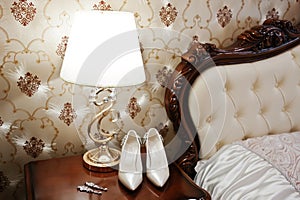 Elegancy wedding shoes of bride with brilliant brooch and earrings on bedside table with desk lamp