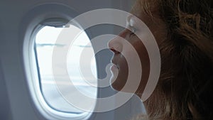 Elegant adult woman is sitting inside a plane, looking in porthole on a clear blue sky and texting messages in her phone