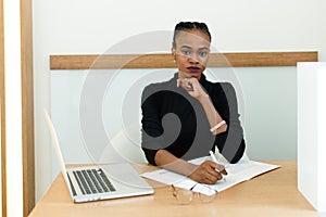 Elegant African or black American woman holding chin working at desk with notepad and laptop in office