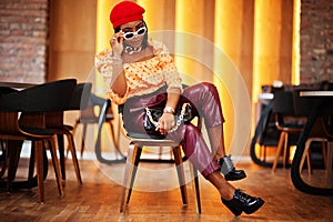 Elegant afro american woman in red french beret, big gold neck chain, sunglasses, polka dot blouse and leather pants pose indoor