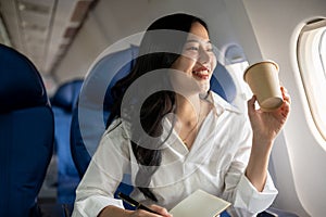 An elegant Asian businesswoman is sipping coffee while looking at the view outside the plane window