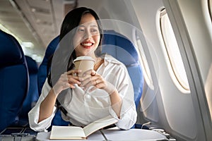 An elegant Asian businesswoman is sipping coffee while looking at the view outside the plane window