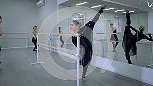 Elegant beautiful ballerina rehearsing arabesque in dance studio with ballet dancers at background. Wide shot portrait