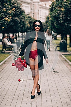 Elegant beautiful brunette woman in red shorts and black coat posing in the alley with a big bouquet of red roses