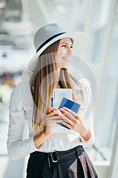 Elegant beautiful business girl holding travel tickets and using phone, happy girl looking at camera in airport terminal