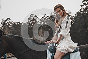 Elegant and beautiful magnificent young woman wearing the dress is holding reins and riding a horse